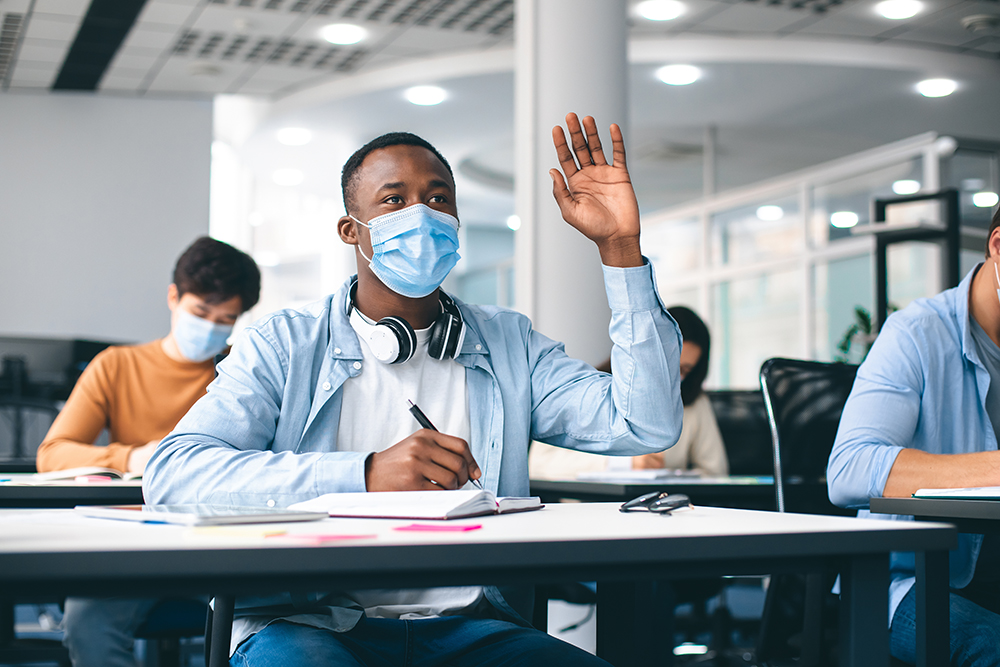 Transferência Graduação: Estudante afro-americano levantando a mão para responder, usando máscara médica protetora para proteção contra doenças virais na aula do ensino médio